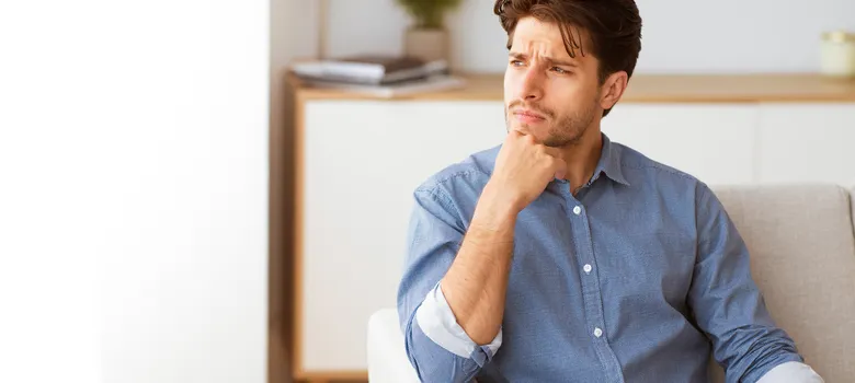 man-wearing-blue-shirt-sits-white-couch-indoors-hand-his-chin-looking-thoughtfully